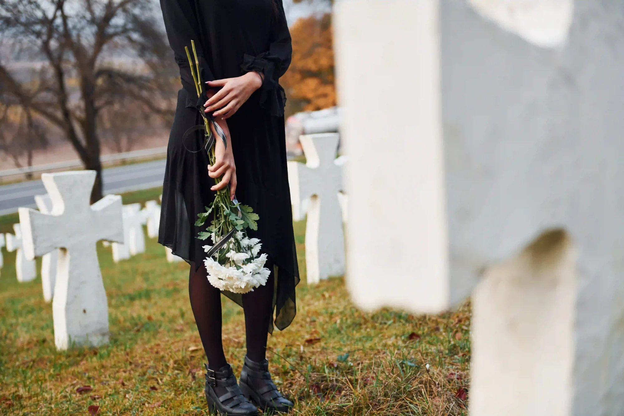 Man sieht eine Frau in schwarzer Trauerkleidung, die einen Strauss weißer Nelken in der Hand hält und auf einem Friedhof mit weissen Keuzen an einem Grab steht.