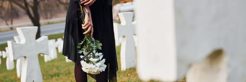 Man sieht eine Frau in schwarzer Trauerkleidung, die einen Strauss weißer Nelken in der Hand hält und auf einem Friedhof mit weissen Keuzen an einem Grab steht.